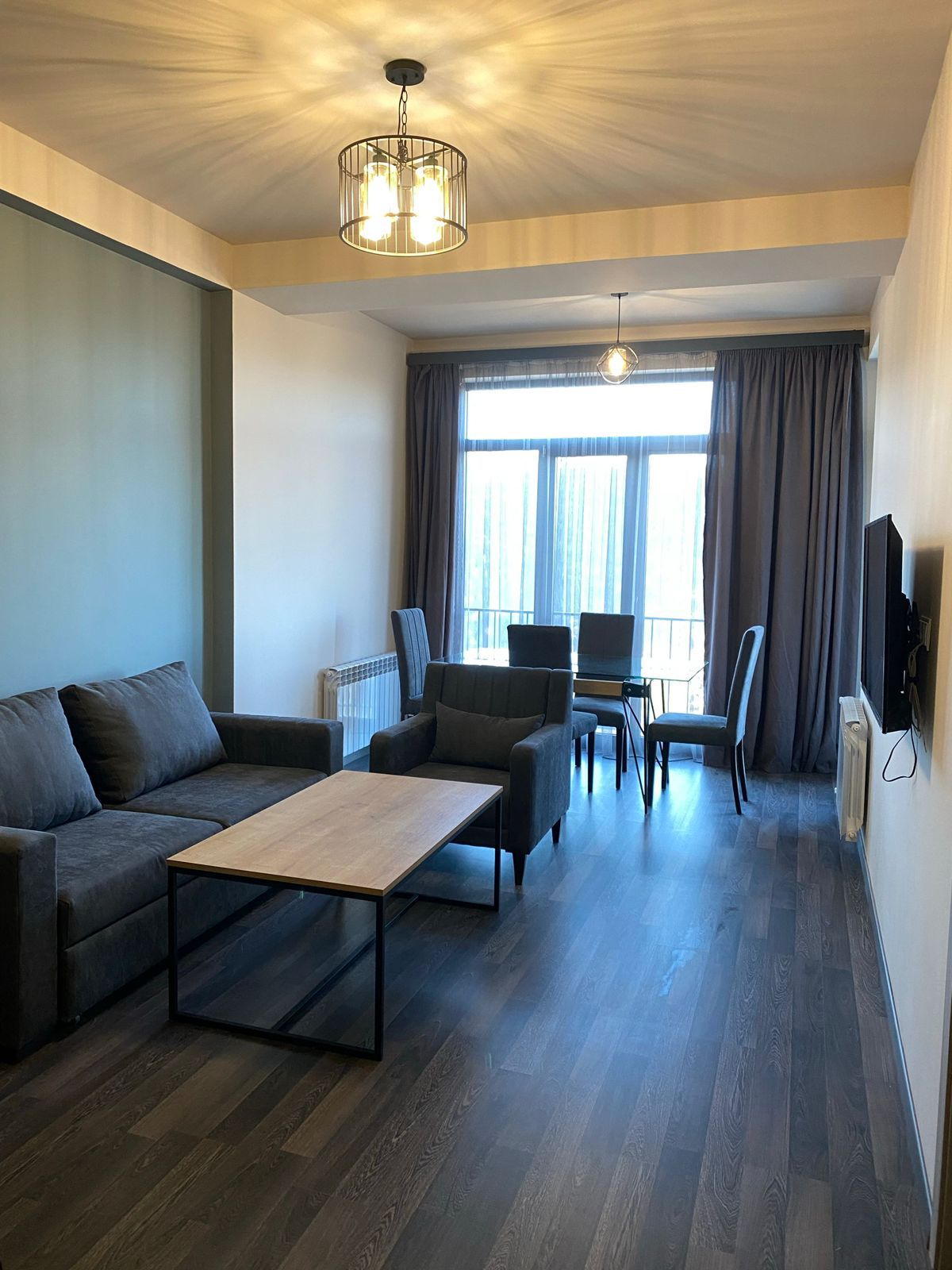 A cozy living space is equipped with a dark gray sofa positioned beside a light wooden coffee table. A dining area is visible with a rectangular table and four chairs. Natural light filters in through large glass doors leading to a balcony, accompanied by soft, neutral-colored drapes.