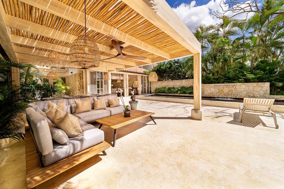 An open-air seating area features a spacious sectional sofa adorned with soft cushions and a wooden coffee table. Natural light filters through the overhead bamboo covering, while palm trees and lush greenery create a serene outdoor backdrop. A sun lounger stands nearby on the stone floor.