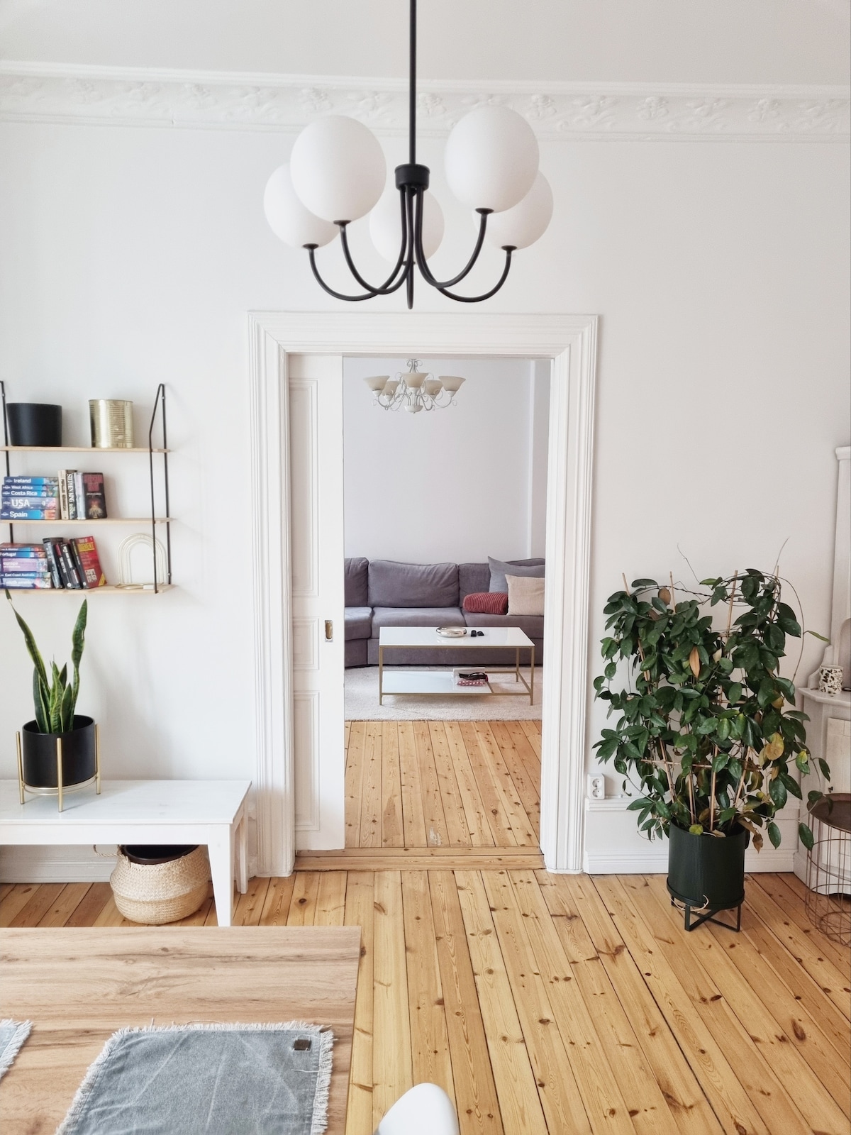 A bright living space is showcased with light-colored walls and a warm wooden floor. A modern chandelier hangs from the ceiling, while a cozy seating area is visible through the doorway. Shelves with books and decorative items are displayed on the wall, alongside potted plants.
