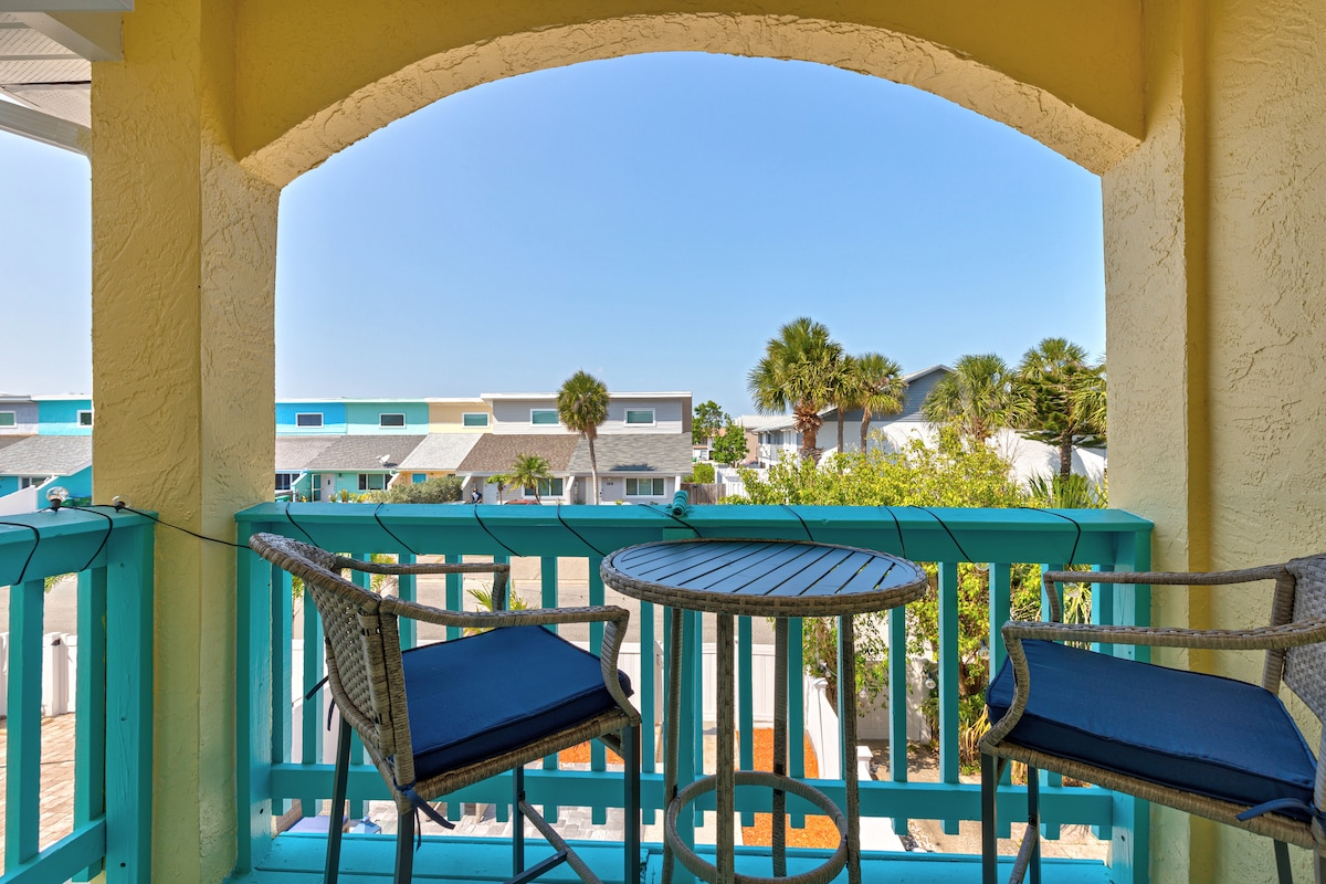 A private balcony is featured with two cushioned chairs and a small round table, providing a view of the surrounding area. The railing is painted in a bright blue, contrasting with the soft yellow walls and showcasing palm trees in the distance.