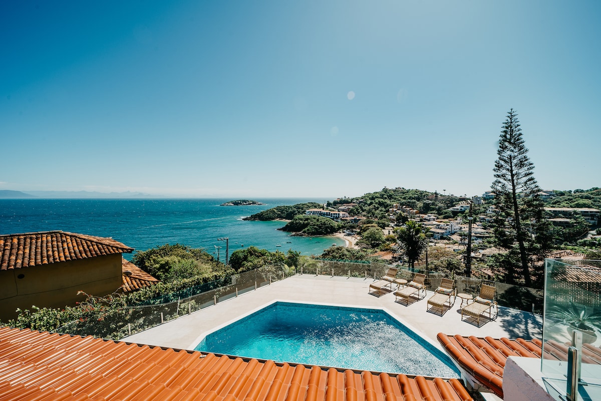 A clear view of a sparkling pool is presented, surrounded by sun loungers. The horizon showcases a scenic coastline with lush greenery and distant houses, under a bright blue sky. The terracotta roof provides a contrasting element in the foreground.