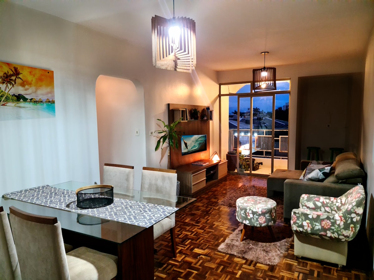The living area features a blend of modern furnishings, including a glass dining table set for six and a cozy seating arrangement with floral-patterned and neutral-colored sofas. A wall-mounted television is visible, while glass doors lead to a balcony, allowing natural light to fill the room.