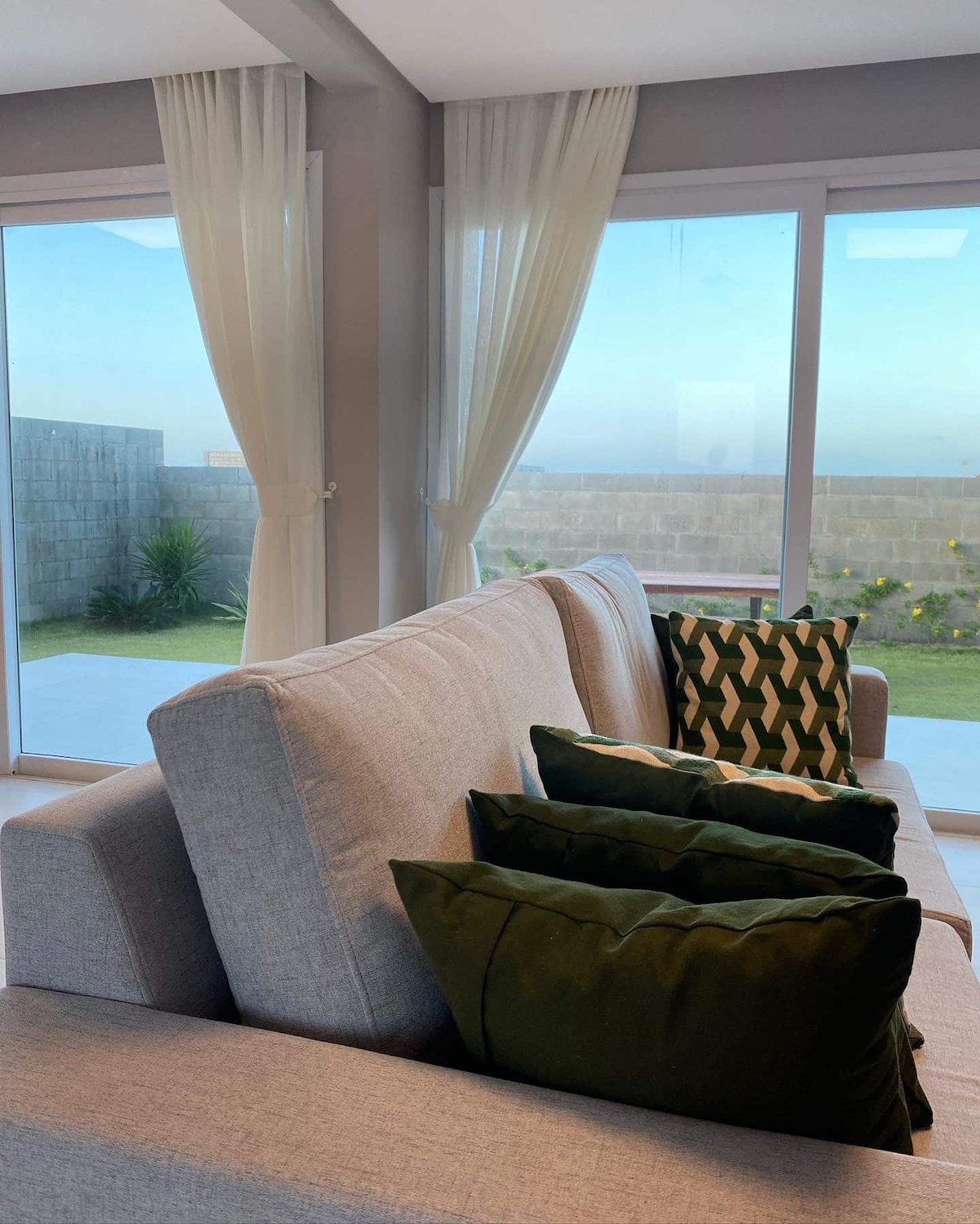 A light-filled living area features a soft gray sofa adorned with patterned and solid green cushions. Large glass doors frame views of the outdoor garden, with sheer curtains gently diffusing natural light, creating an airy ambiance.