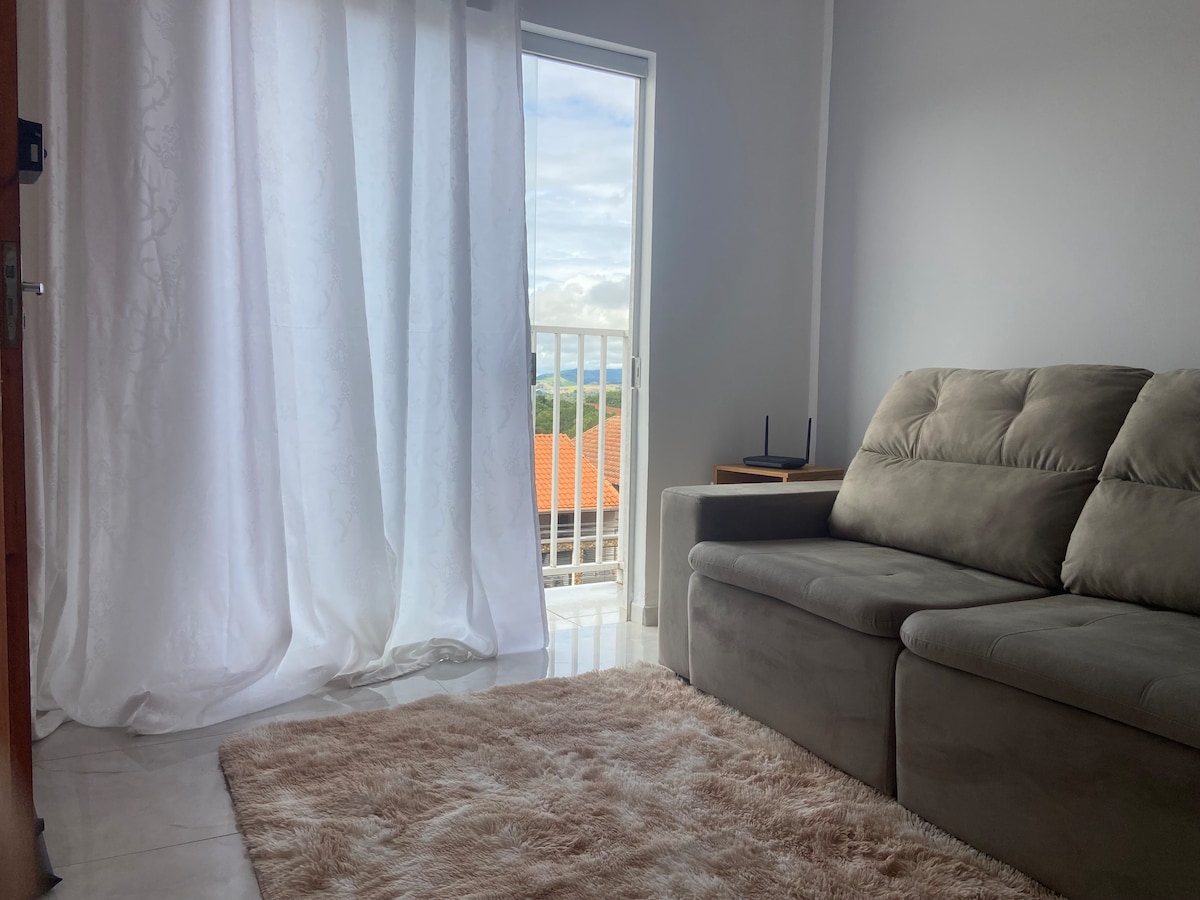 A bright living area features a comfortable gray sofa positioned near a large window. Sheer white curtains filter natural light and frame the view, while a soft area rug adds warmth to the tiled floor.