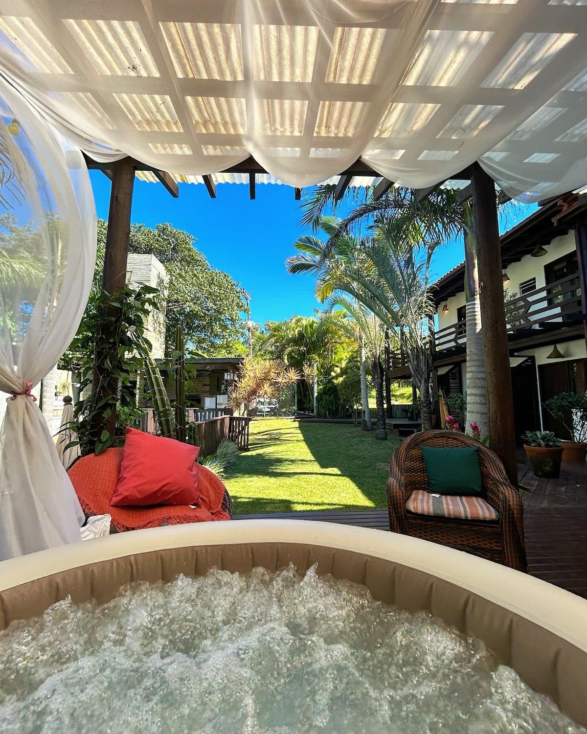 A relaxing outdoor area features a hot tub surrounded by lush greenery and palm trees. The gazebo is adorned with sheer curtains, providing shade and creating a serene atmosphere. Bright accent cushions are placed on nearby seating, enhancing the space's comfort and leisure appeal.