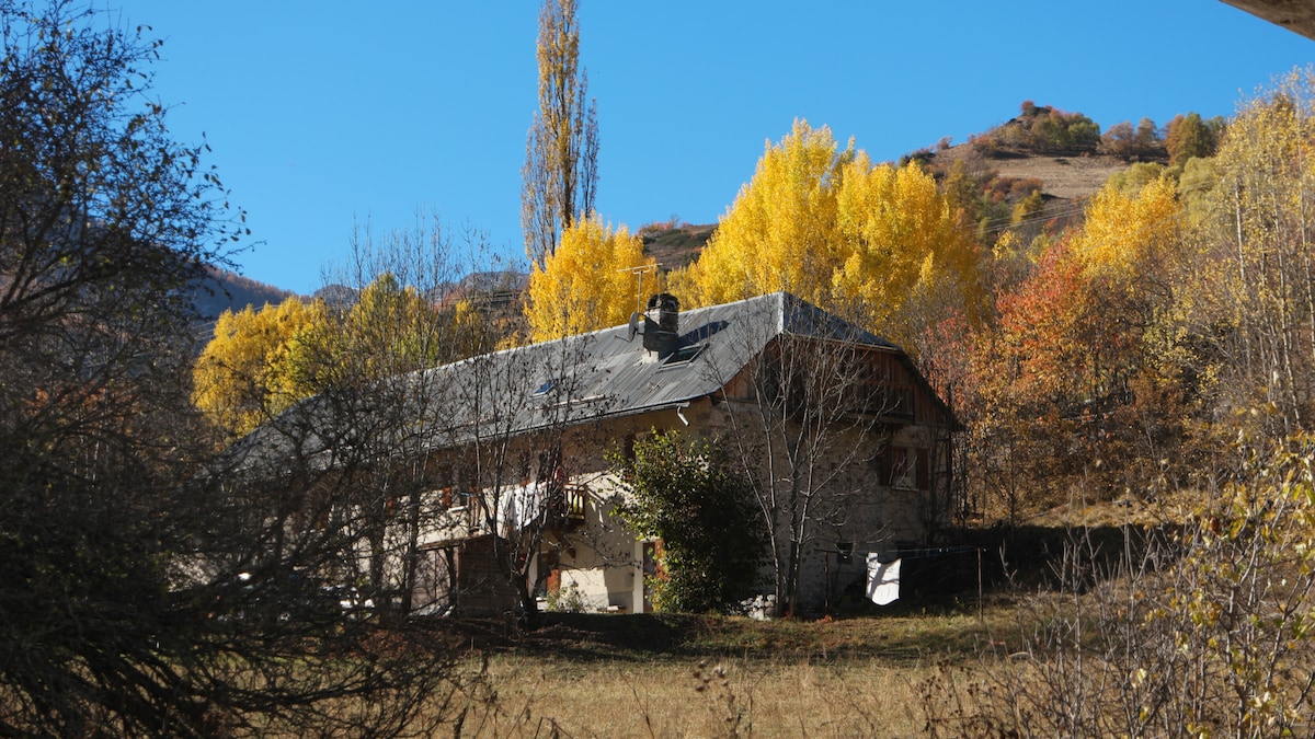 A rustic mountain house is surrounded by autumn foliage, showcasing vibrant yellow and orange trees. The stone walls and sloped roof of the house reflect the natural landscape, while a clear blue sky serves as a backdrop.