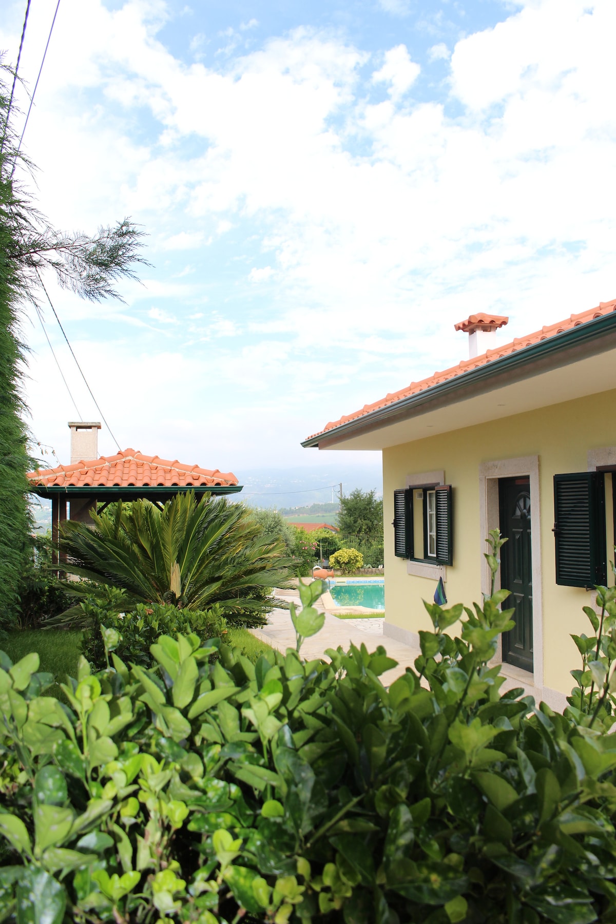 The image presents a charming yellow house situated amidst lush greenery and tropical plants. A bright red tiled roof contrasts with the green shutters on the windows. A glimpse of the private pool can be seen in the background, under a partly cloudy sky.