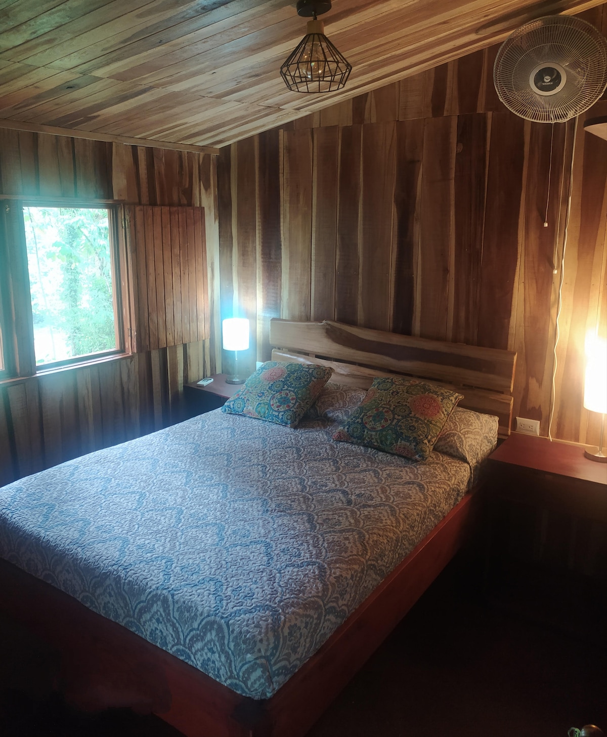 A cozy sleeping area is presented with wooden paneling on the walls, creating a natural ambiance. A spacious bed is adorned with a patterned blanket and two decorative pillows, complemented by bedside lamps. Natural light enters through a window partially draped with wooden shutters.