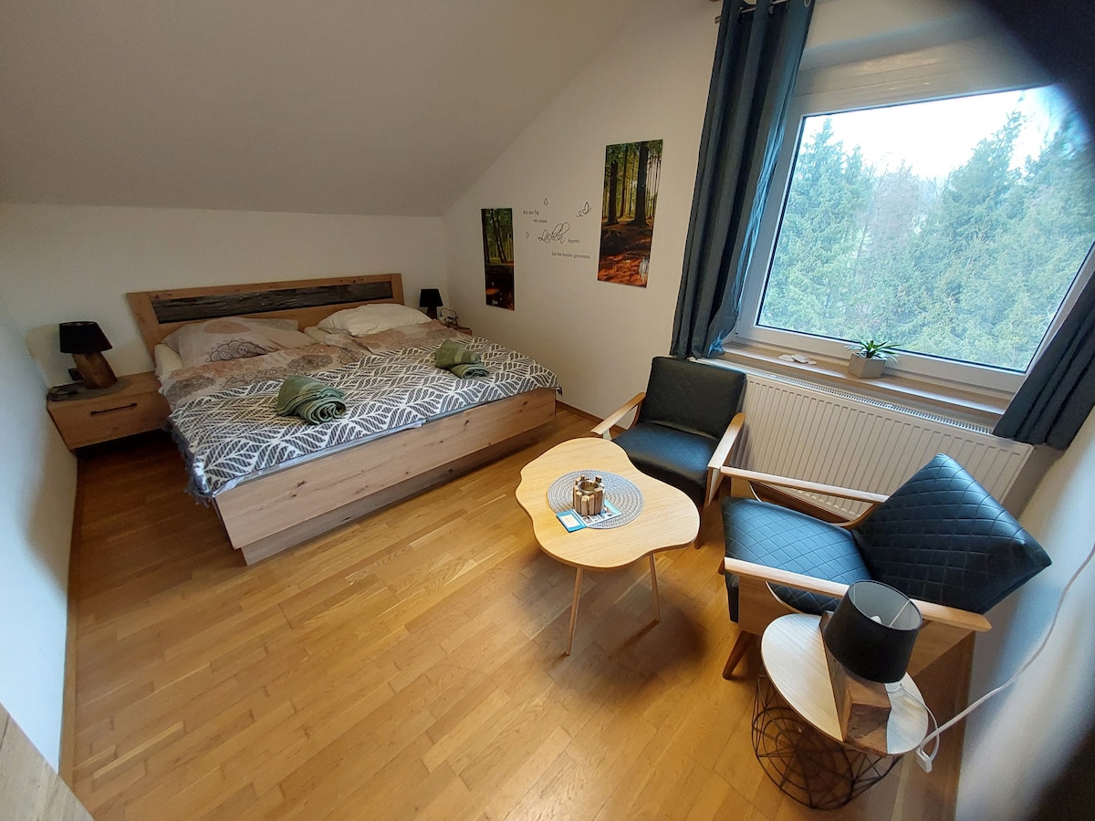 A spacious bedroom features a double bed with a textured blanket, two bedside tables, and lamps on either side. A small seating area with two chairs and a coffee table is positioned near a large window, allowing natural light to illuminate the wooden floors.