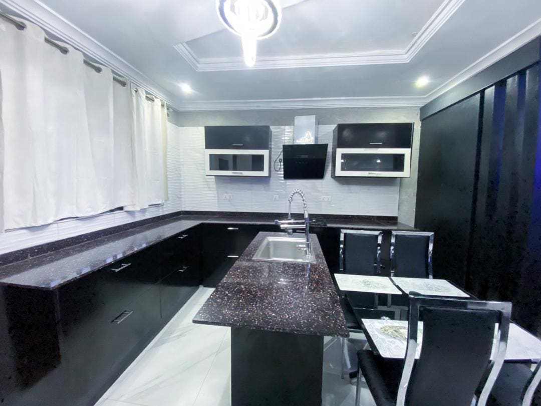 A modern kitchen is presented with sleek black cabinetry and a glossy countertop. A sink is situated on an island with seating for four. Upper storage cabinets are equipped with glass doors, and a flat-screen TV is mounted on the wall. Soft lighting enhances the space.