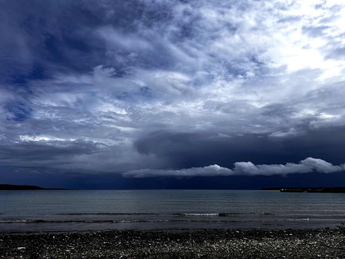A tranquil waterfront view is presented, featuring a calm lake under a dramatic sky filled with various shades of gray and blue. Soft ripples are visible on the water's surface, reflecting the cloud formations above. The shoreline is composed of pebbles and smooth stones.
