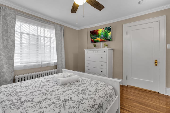 A large bedroom with a queen-sized bed, Sonos smart speaker, a Roku TV, chest, and phone chargers.
