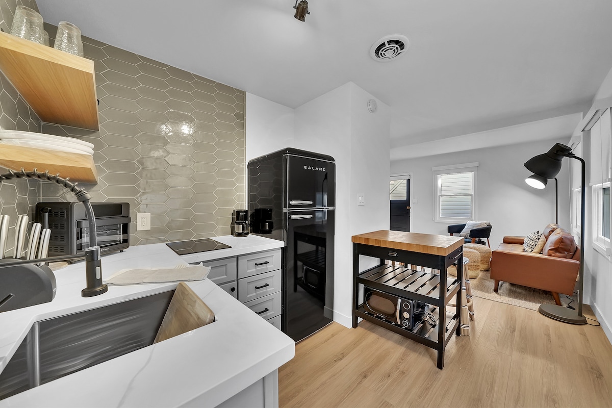A modern kitchen area features sleek cabinetry and a retro black refrigerator. Open shelving displays dishware, while a large butcher block island provides ample workspace. The adjacent living area is visible, showcasing comfortable seating and natural light filtering through the windows.