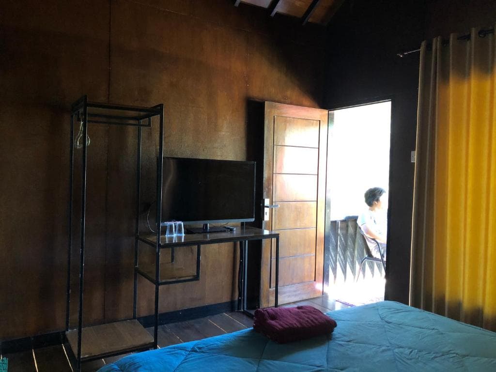 A cozy room is shown with wooden walls and a spacious bed covered in a turquoise quilt. A small TV is mounted on a stand. Natural light filters through yellow curtains, illuminating the entrance door that leads to a terrace.