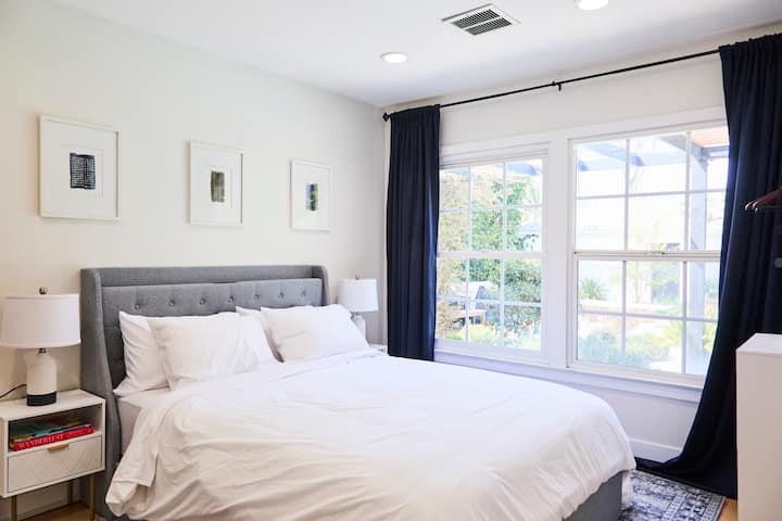 This serene bedroom features a plush gray tufted headboard, crisp white bedding, and matching nightstands with modern lamps. Large windows with dark drapes bring in natural light while allowing for cozy privacy.
