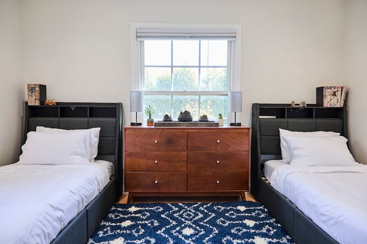 The twin bedroom offers two cozy beds with crisp white linens, built-in headboard shelving, and a spacious dresser. A bold patterned rug adds style, while the sunny window brightens the space—perfect for kids, friends, or extra guests.
