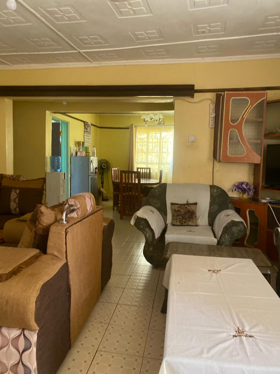 A spacious living area is presented with comfortable seating options, including a mix of sofas and chairs. A coffee table occupies the center, while a dining area is visible in the background. Natural light enters through a window, illuminating the warm-colored walls.