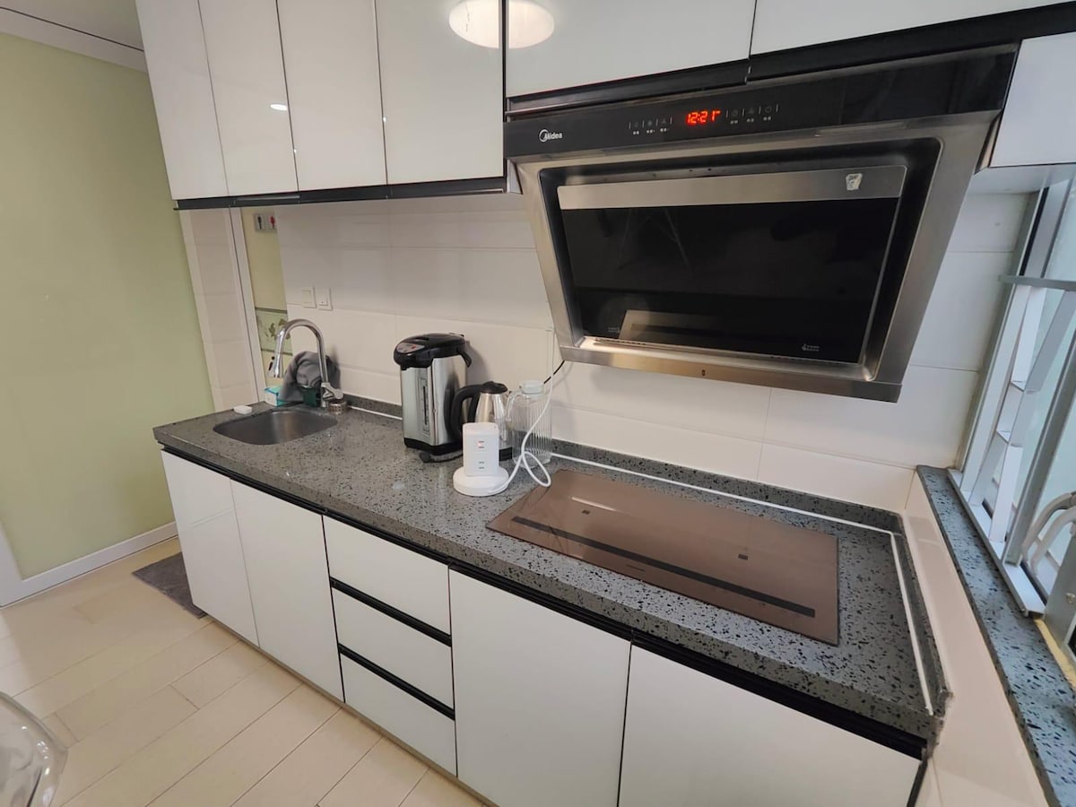A modern kitchen features sleek white cabinetry and a grey speckled countertop. A stainless steel kettle and a sleek microwave are prominently displayed. The kitchen layout includes an under-mount sink and a built-in stovetop, with natural light streaming through a nearby window.