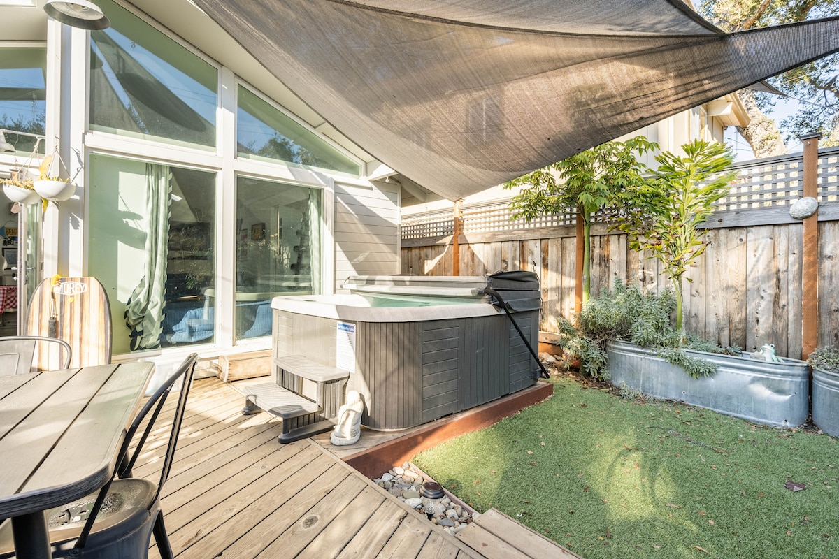 A private outdoor space features a hot tub surrounded by greenery and wooden decking. A shaded area protects the hot tub, while a dining table with chairs is positioned nearby. Sunlight filters through the trees, creating a peaceful atmosphere.