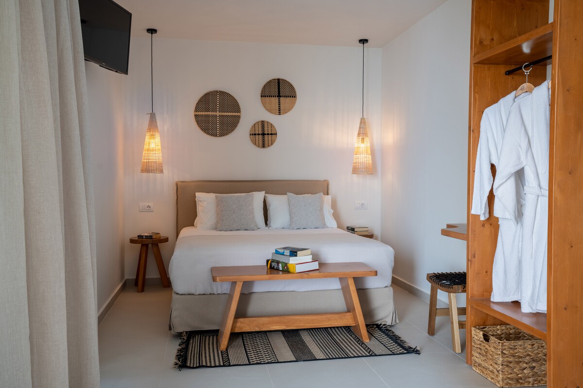 A spacious bedroom features a comfortable queen-size bed with soft linens, framed by warm wooden decor. Two stylish pendant lights hang above, illuminating the space. Natural light streams in, complemented by a small seating area and open wardrobe, enhancing the calm ambiance.