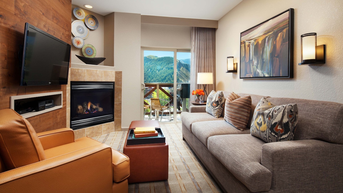 A comfortable living area is depicted, featuring a plush gray sofa adorned with decorative cushions. A leather armchair and a wooden coffee table complement the space. A cozy fireplace is positioned beneath a flat-screen TV, with a balcony offering scenic mountain views in the background.
