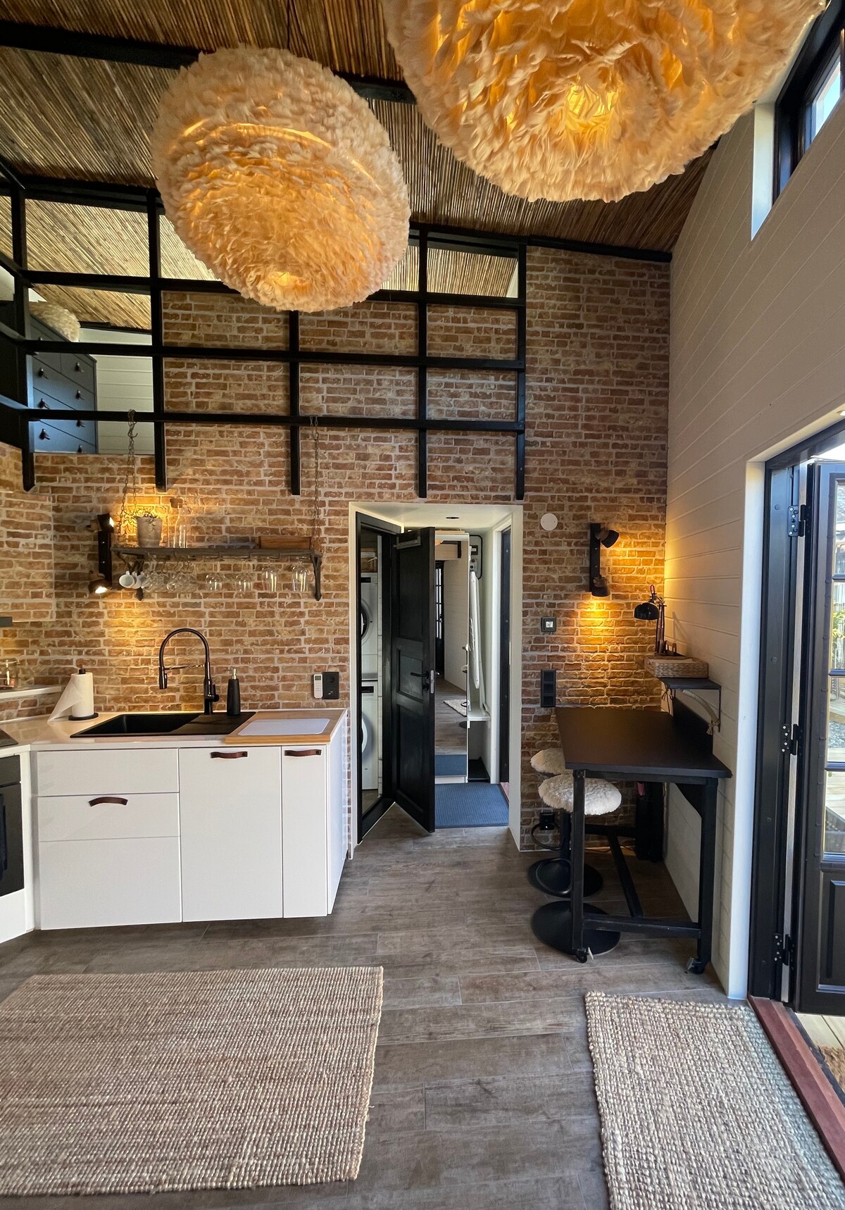 The kitchen area features exposed brick walls and modern cabinetry, complemented by stylish pendant lighting. A black dining table with two stools is positioned beside the kitchen counter, while a small rug adds warmth to the space. Glass doors lead to the outdoor area.