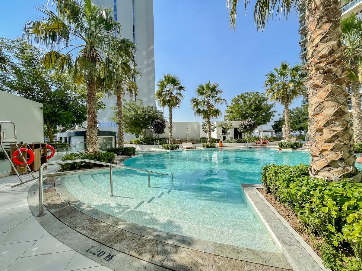 A serene pool area is surrounded by palm trees and well-maintained landscaping. The water is calm, reflecting the clear blue sky above. Lounge chairs are positioned around the pool, offering space for relaxation and enjoyment.
