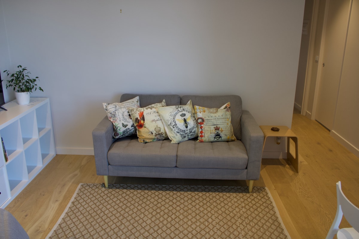 A gray sofa with decorative pillows is positioned against a neutral wall. A small table is placed beside the sofa, and a light-colored area rug adds texture to the hardwood floor. Indoor plants provide a touch of greenery in the corner.