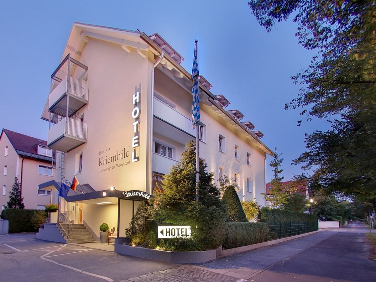 A modern three-story hotel building is depicted, featuring a welcoming entrance with a canopy and well-kept greenery. The façade is accentuated by large windows and balconies on the upper floors. Soft evening light highlights the hotel's name, creating a calming ambiance.