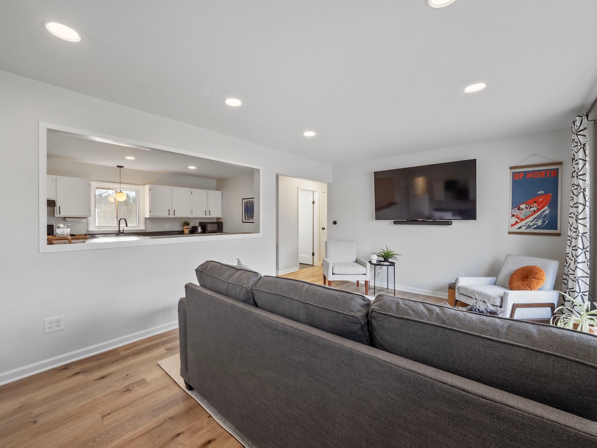 A spacious living area features a comfortable grey sectional sofa facing a large flat-screen television mounted on the wall. Natural light filters in through a window, illuminating the semi-open kitchen visible through a pass-through window.