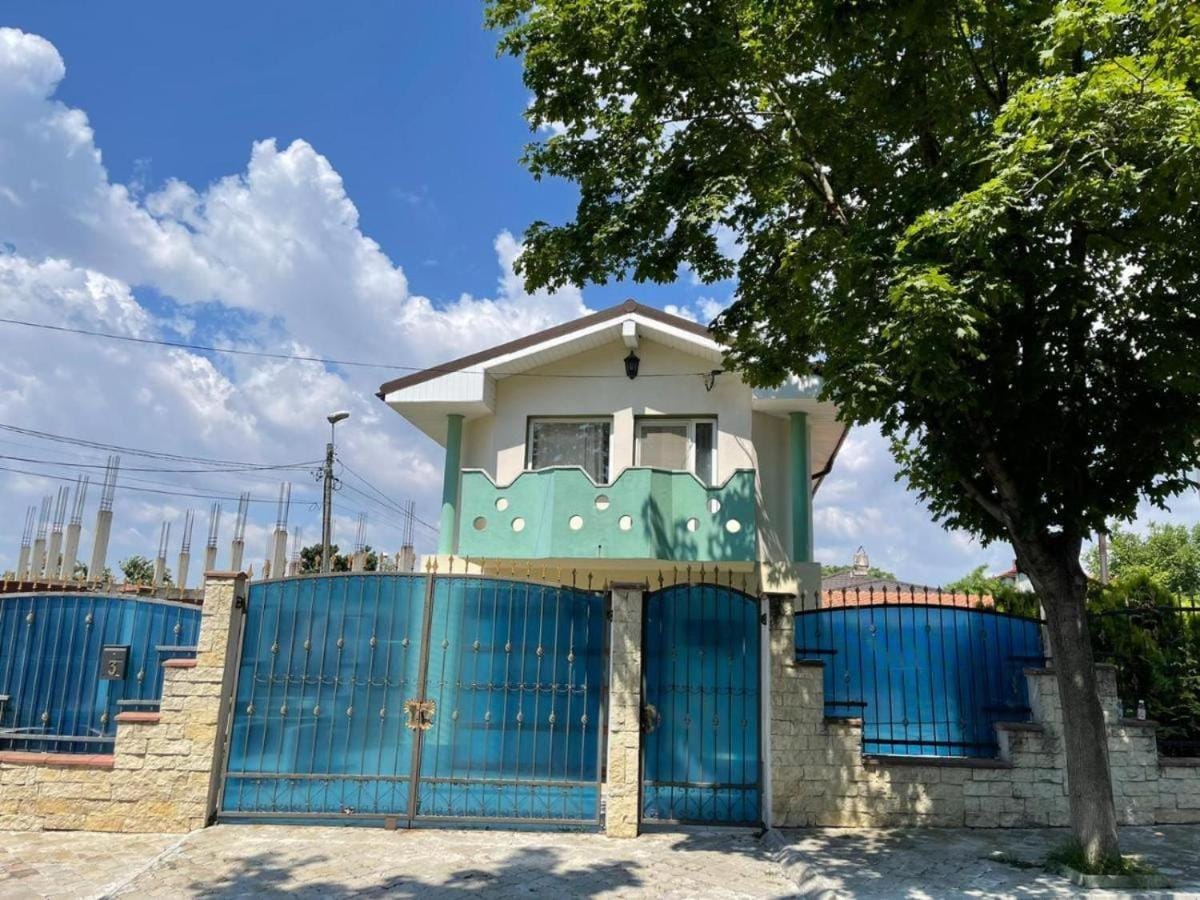 A charming two-story house is framed by lush greenery and features vibrant blue gates. The exterior is adorned with a playful turquoise design, and large windows provide a glimpse of the interior. Soft clouds drift across the sky, creating a calm atmosphere around the property.