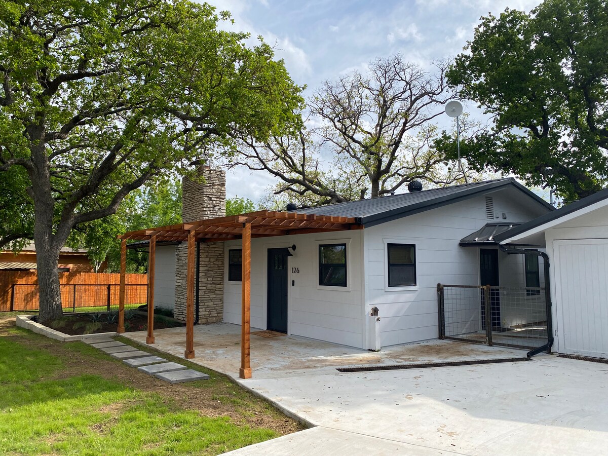 A modern midcentury home is presented with a covered porch supported by wooden beams. A stone chimney enhances the exterior, which features a mix of light-colored siding and large windows. The property is surrounded by mature trees and a grassy yard.