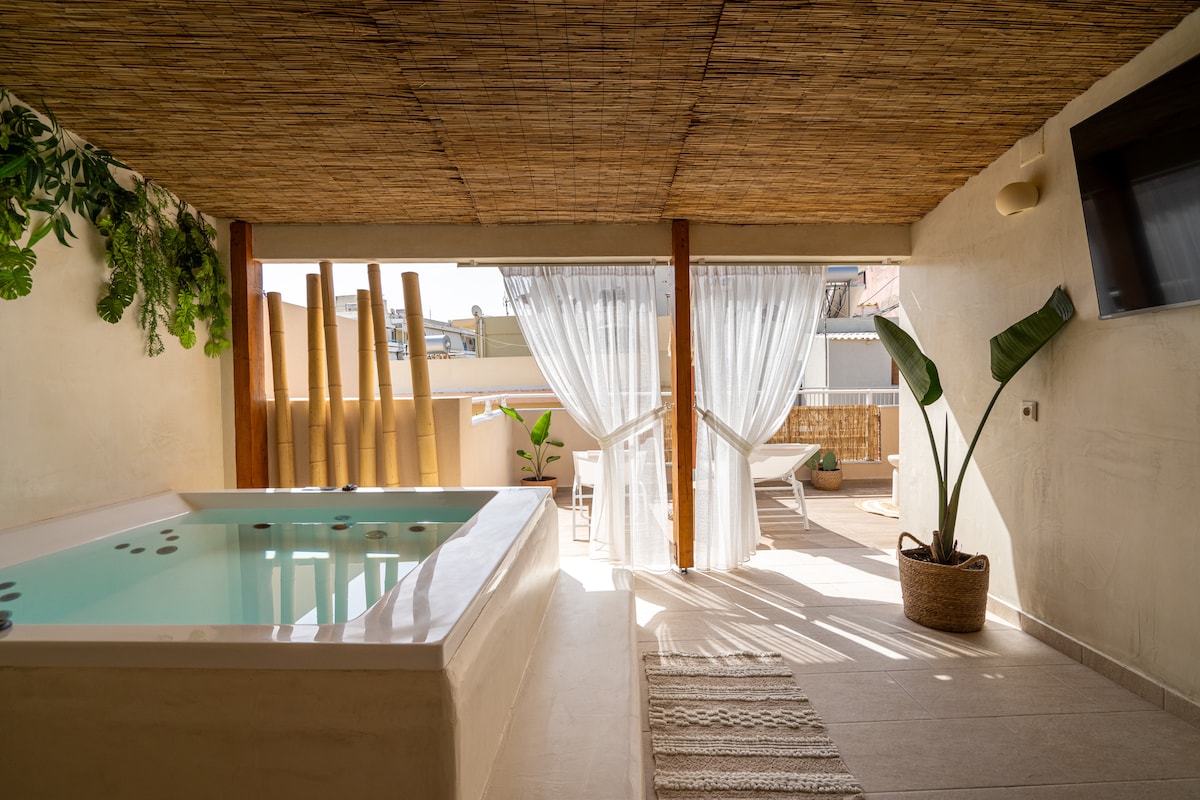 An outdoor area features a private jacuzzi surrounded by bamboo elements and lush greenery. Sheer curtains drape softly, allowing natural light to filter in. A comfortable lounge area with sun loungers is visible under a thatched roof, enhancing the space's relaxed vibe.