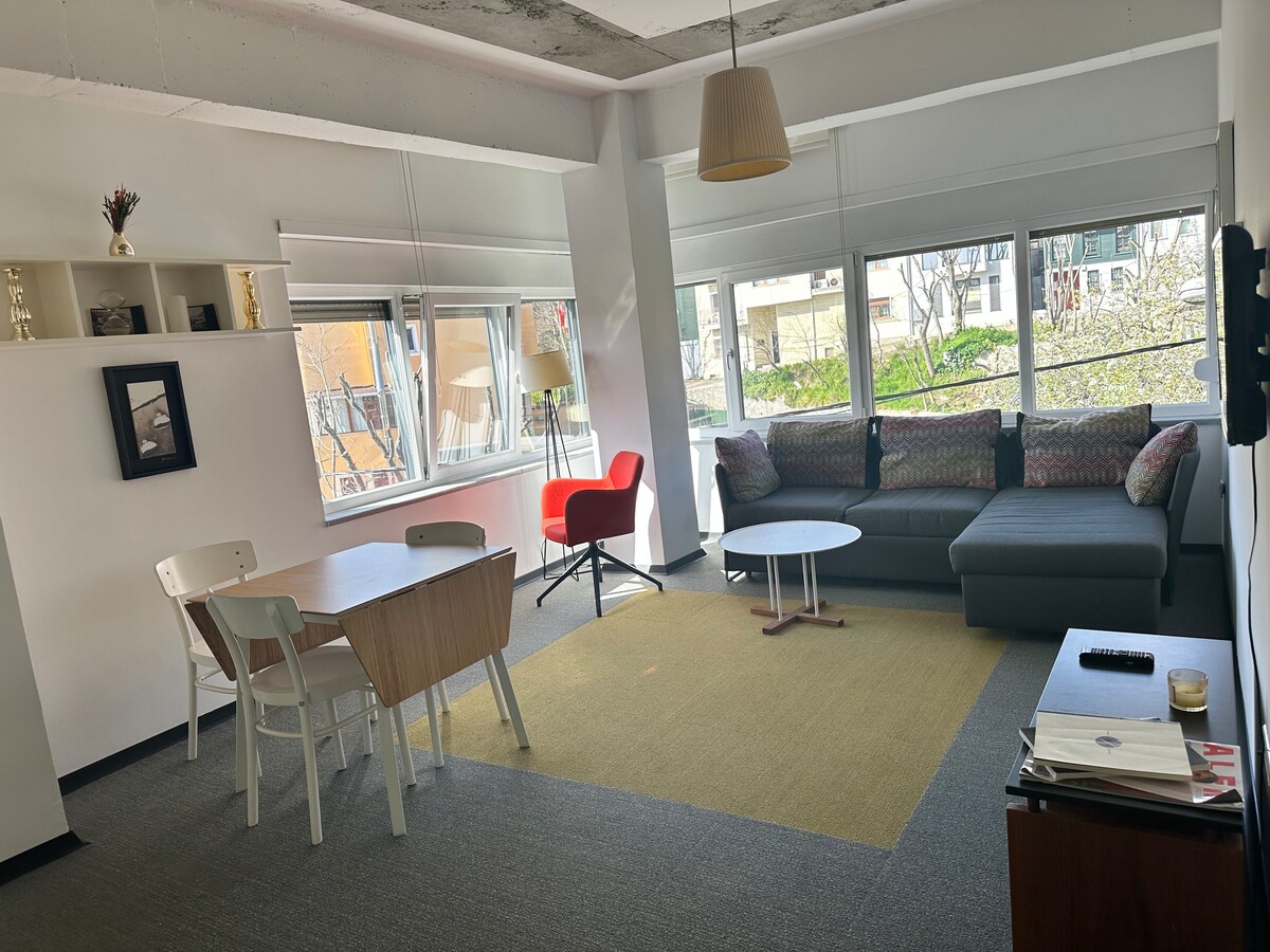 The living area features a spacious sofa with patterned cushions, a round coffee table, and a dining table with three chairs. Large windows allow natural light to fill the room, providing views of greenery outside. A single pendant light hangs from the ceiling, adding to the modern decor.