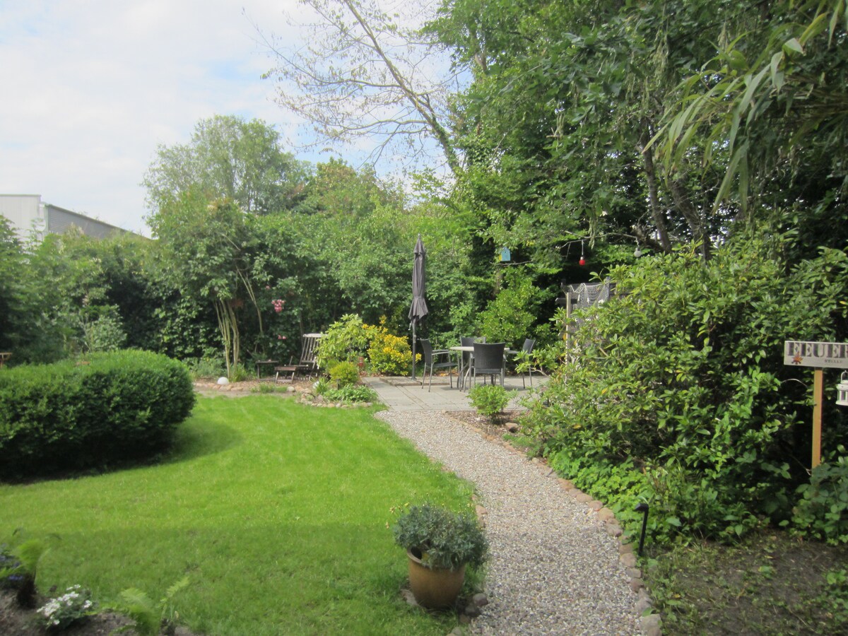 A well-maintained garden is featured, with a winding gravel path leading through lush greenery. Various seating areas, including a table and chairs, are visible amidst the vibrant plants and trees. A sense of tranquility is evoked by the surrounding natural elements.