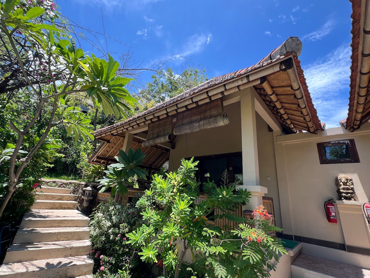 A traditional Balinese-style building is framed by lush greenery, featuring a sloped tiled roof and wooden accents. Steps lead to the entrance, and vibrant plants add color to the surroundings, creating a welcoming entrance in bright daylight.