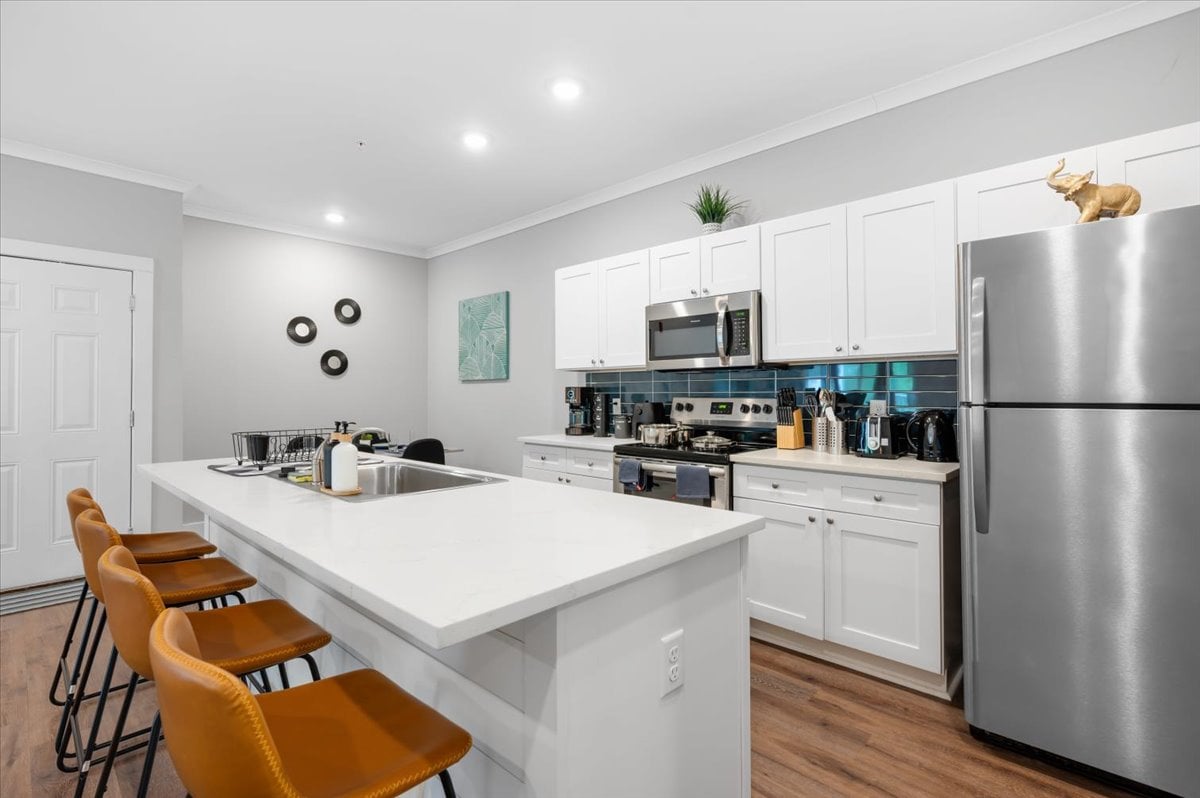 The kitchen features modern appliances and ample countertop space. Bar stools are arranged at the island, providing a casual dining area. The cabinetry is white, complemented by a sleek, stainless steel refrigerator. Decorative items and artwork add a touch of style to the space.