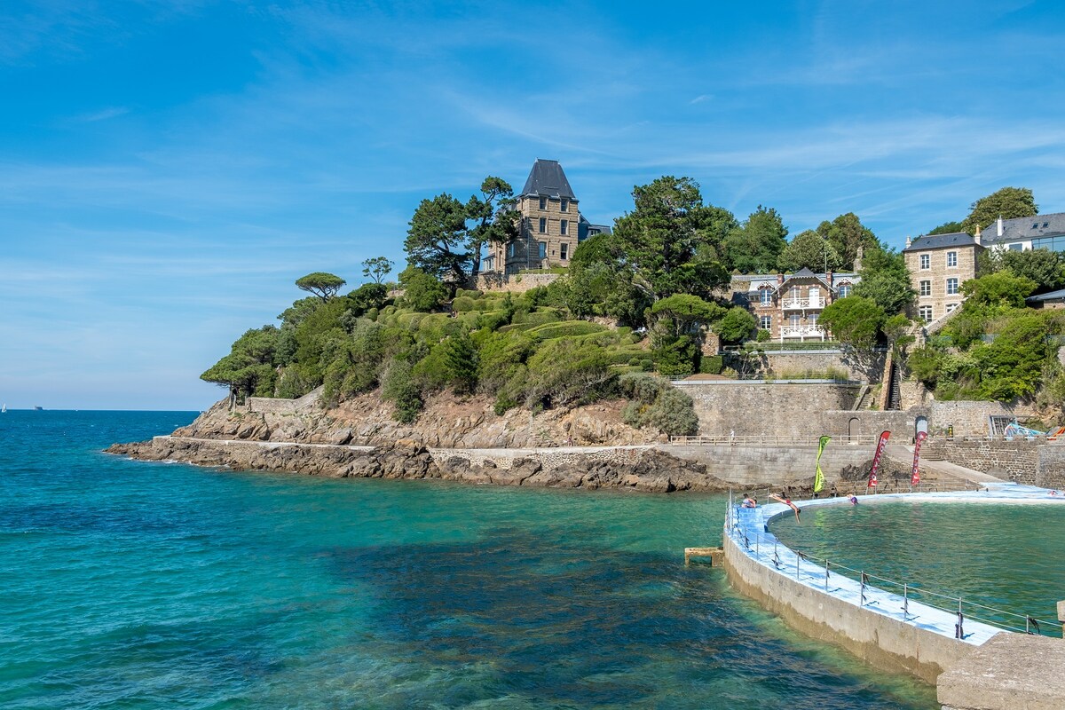 A coastal view showcases the rocky shoreline and clear blue waters, with lush greenery leading to a charming building atop the hill. A public swimming area is visible in the foreground, framed by a circular fence, inviting quiet leisure and scenic enjoyment.