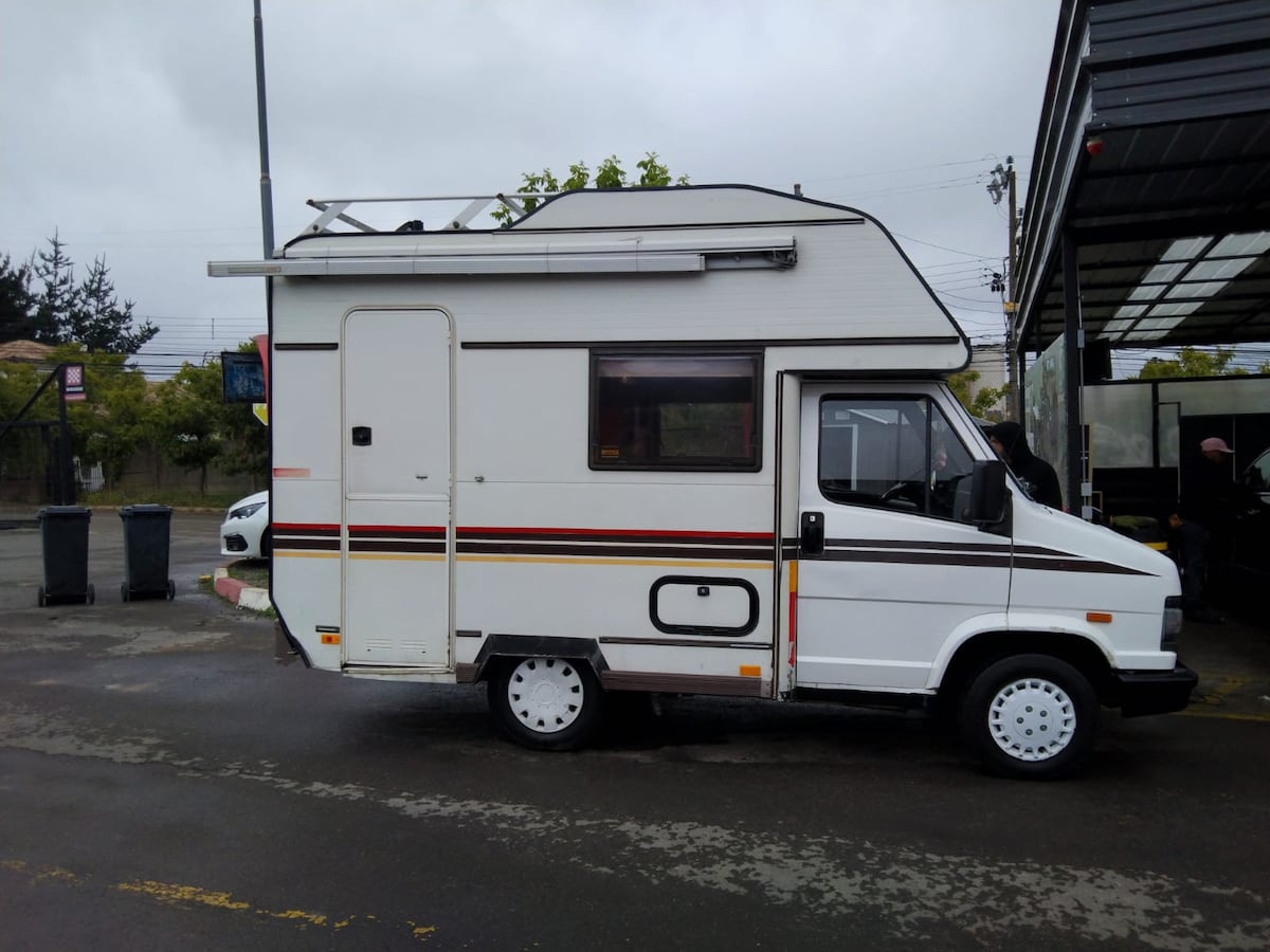 A vintage camper van is parked on a paved surface, showcasing its streamlined body and classic design. The exterior is primarily white with subtle stripes along the sides. An extended roof overhang is visible, along with a side window and a rear awning.