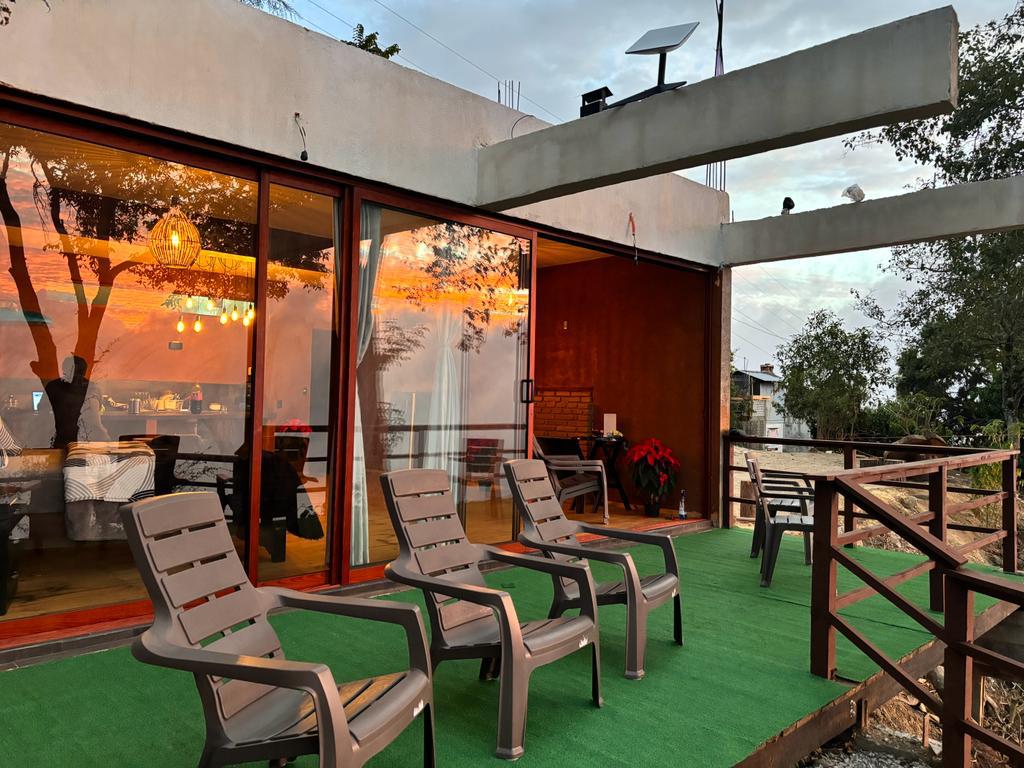 A covered terrace features several lounge chairs arranged on green flooring. Large glass doors provide a view of the interior, while trees and open spaces create a natural backdrop. The sunset casts a warm glow, reflecting off the glass surfaces.