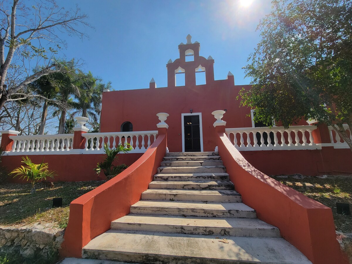 A striking facade of a red-hued building is presented, featuring decorative architectural elements at the top. Wide stone steps lead toward the front door, flanked by white railings and lush greenery that adds a touch of nature to the entrance.