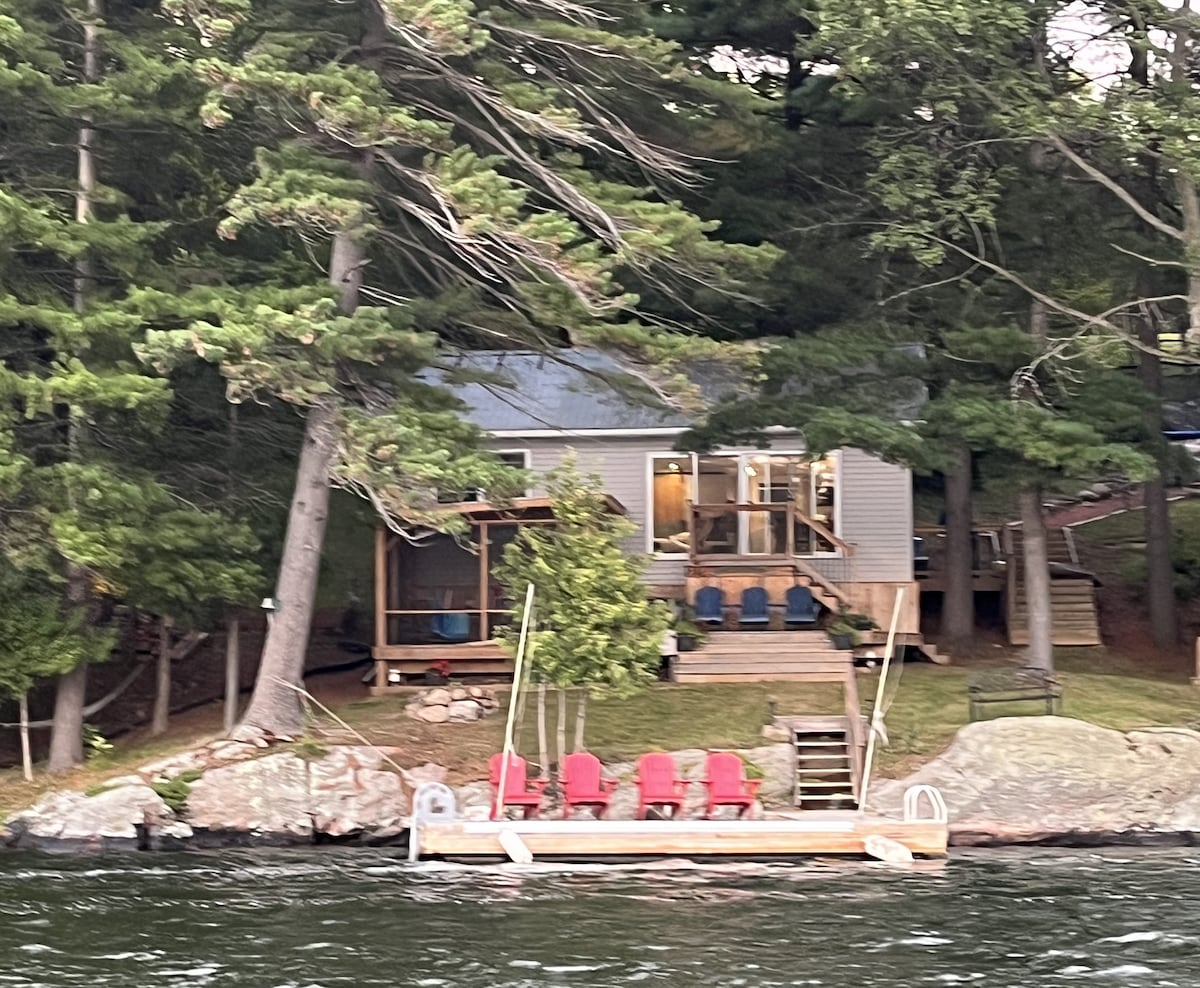 A private cottage is set along the waterfront, surrounded by tall trees. A large floating dock with several red chairs provides a comfortable seating area. The open-concept living space features large windows, allowing for ample natural light and lake views.