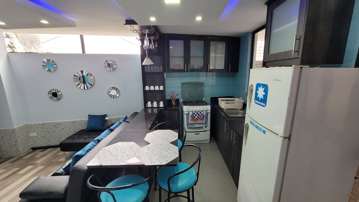 A modern kitchen area features a sleek design, with a dark cabinetry complemented by light-colored countertops. Dining is offered at a geometric table surrounded by four blue chairs. A refrigerator is visible alongside a gas stove, while decorative mirrors adorn the wall.