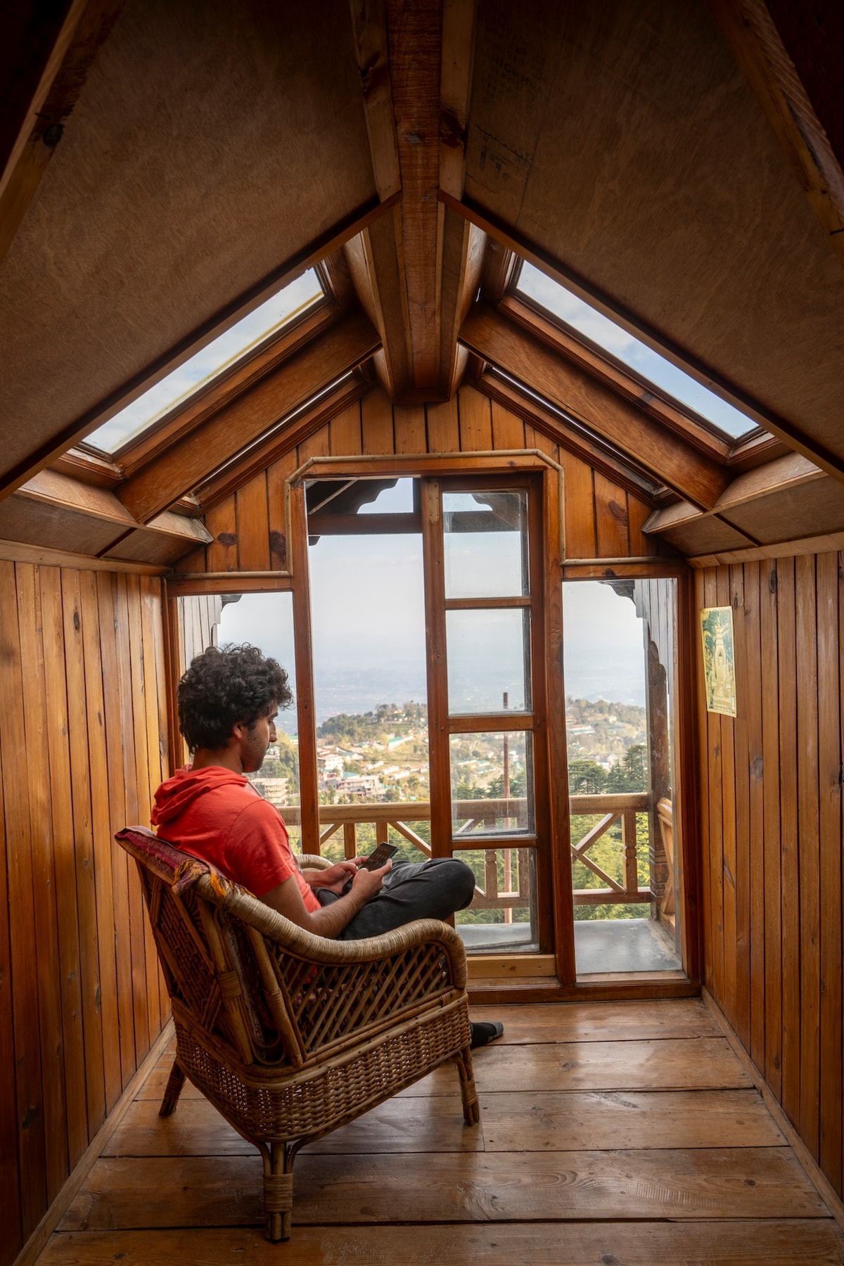A cozy seating area is framed by wooden walls and a sloped ceiling, featuring multiple skylights. A person sits comfortably in a woven chair, enjoying a view through an open glass door that leads to a scenic outdoor landscape.