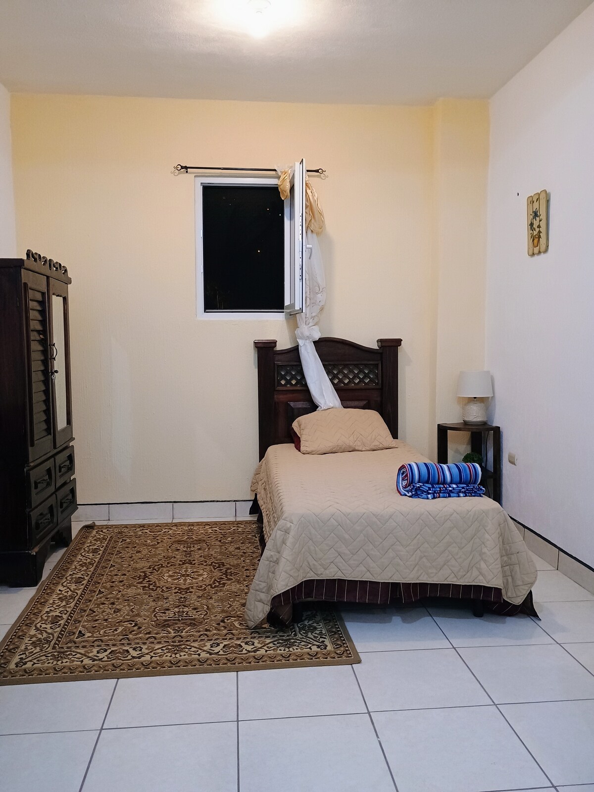 A simple sleeping area features a wooden bed adorned with a light-colored quilt. A striped towel is neatly placed on the bed. A wooden dresser stands against the wall, and a small lamp is positioned beside the bed. Natural light enters through a partially open window.