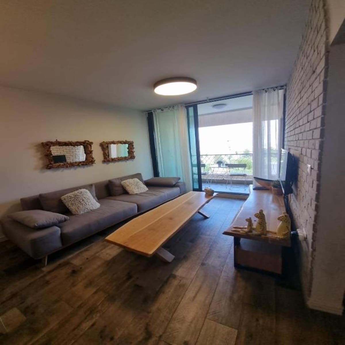 A living area is presented with light-colored walls and wooden flooring. A spacious gray sofa is positioned across from a wooden coffee table, with decorative elements placed atop. Two mirrors framed in natural wood adorn the wall, while large glass doors open to a balcony offering views.