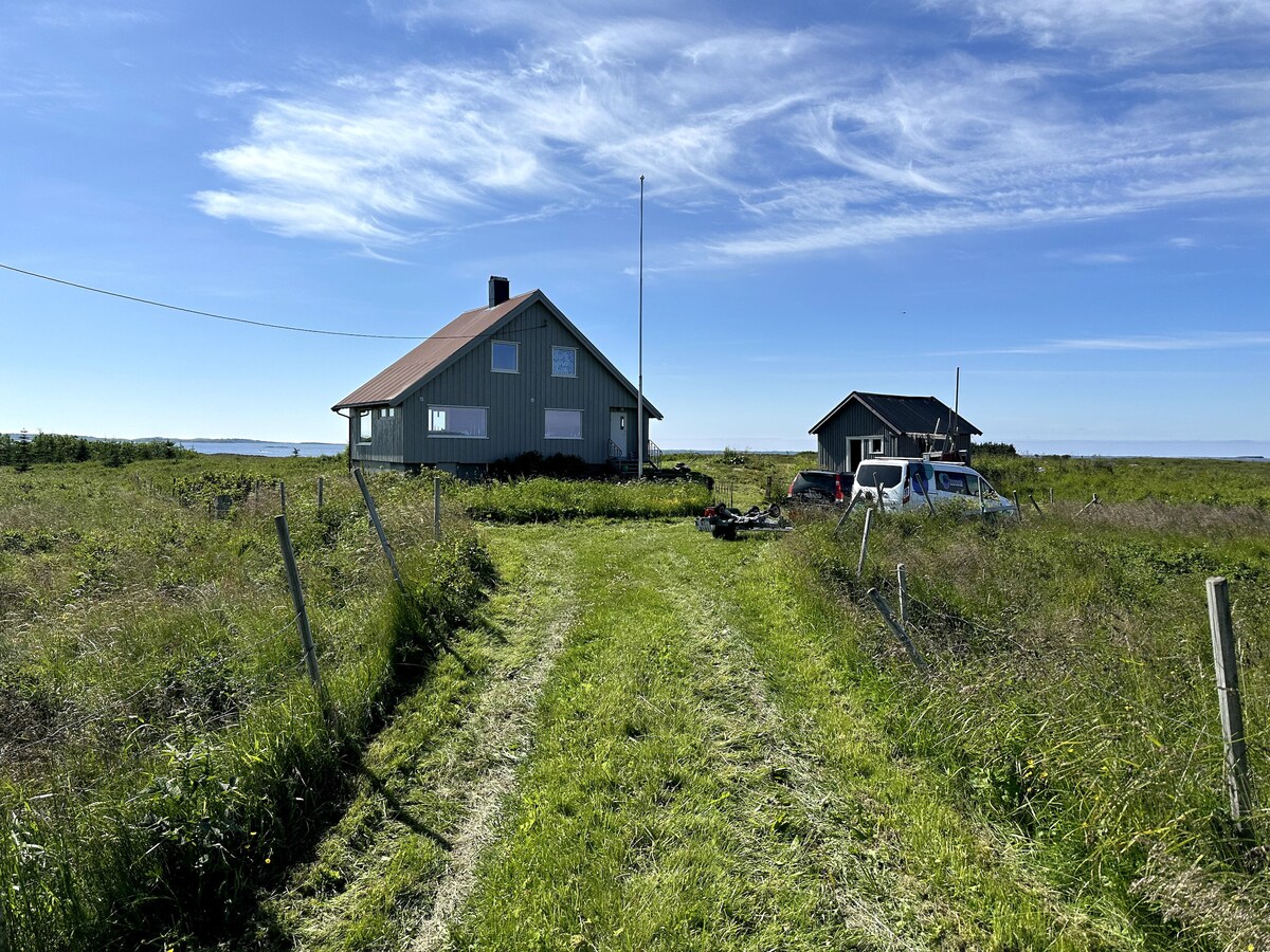 A grassy pathway leads to a two-story house with a red roof, surrounded by open fields under a clear blue sky. A second smaller building is visible to the right, with a parked vehicle nearby. The serene rural landscape conveys a sense of tranquility.