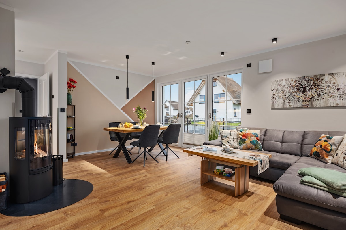 The living area features a comfortable sectional sofa adorned with decorative pillows, a modern coffee table, and a dining area with stylish chairs. Large glass doors allow natural light and views of the outdoor space. A sleek wood-burning stove is positioned nearby.