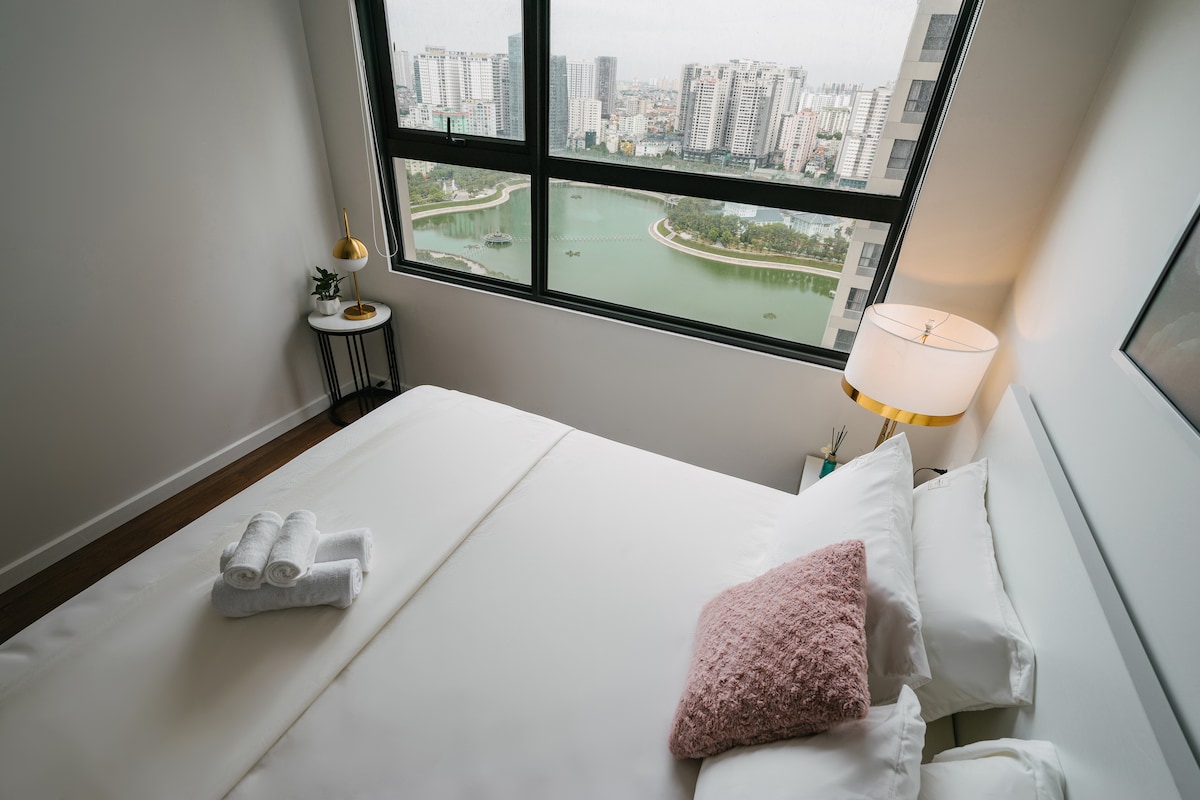 A modern bedroom is showcased, featuring a spacious bed with soft pillows and neatly arranged towels. A large window offers a panoramic view of the city and lake, allowing for ample natural light to fill the room.