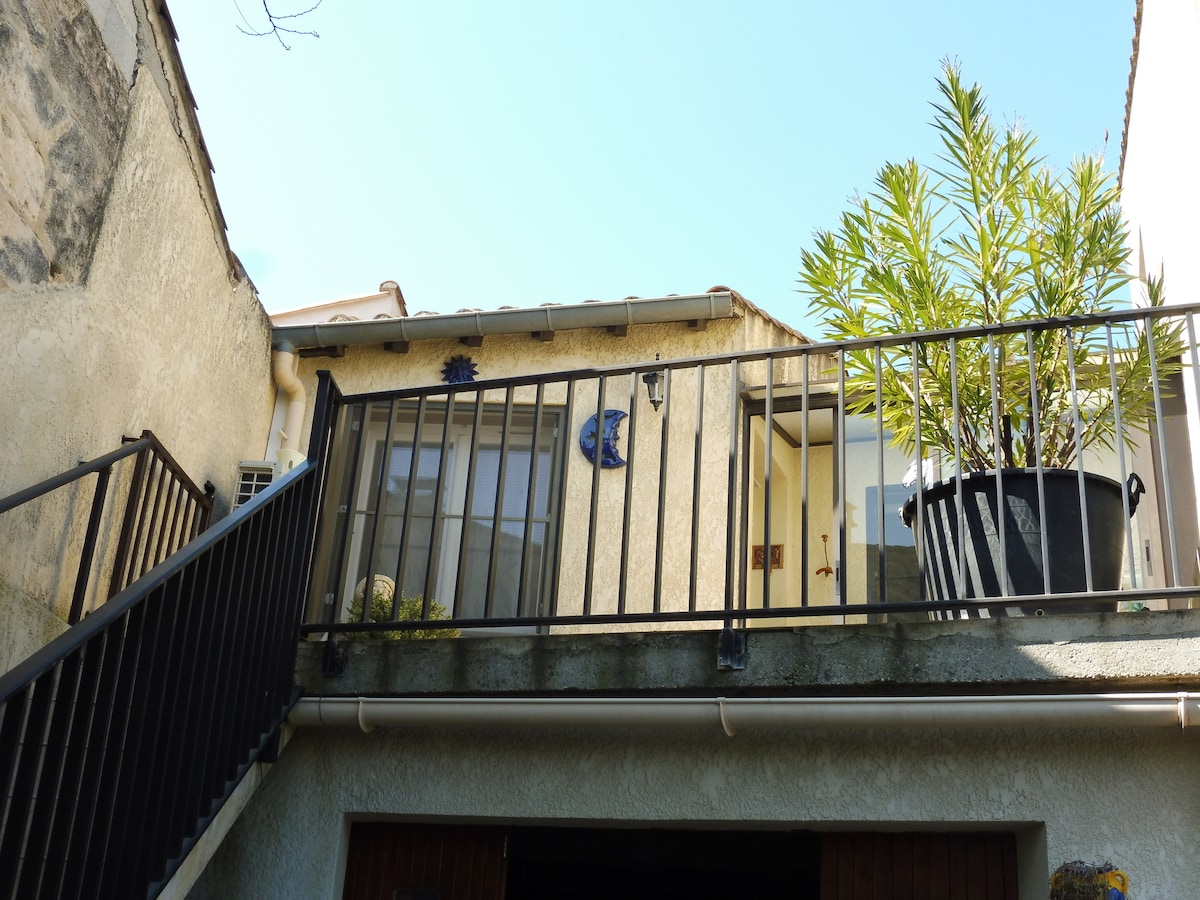A staircase leads to an upper deck, featuring a railing and a large potted plant. The exterior wall displays a light color, with decorative elements visible near the entrance.